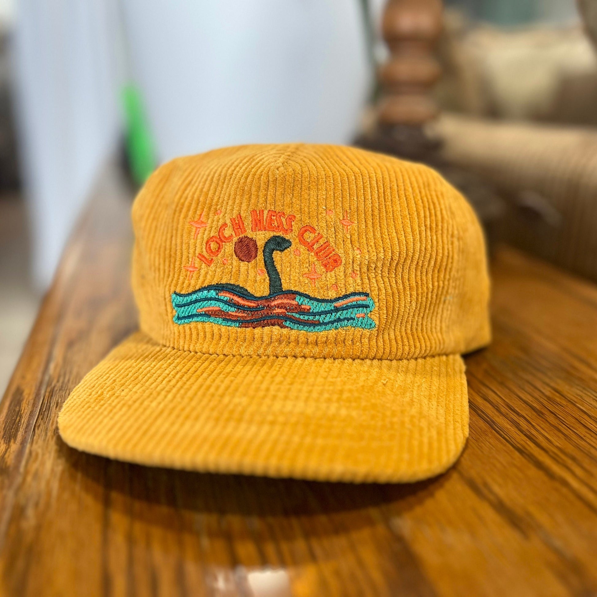 A mustard yellow hat, with direct embroidery depicting the Loch Ness Monster emerging from the water in a silhouette and minimalist style. Features the text "Loch Ness Club" arched above the image. Colors are greens, blues, and warm tones.
