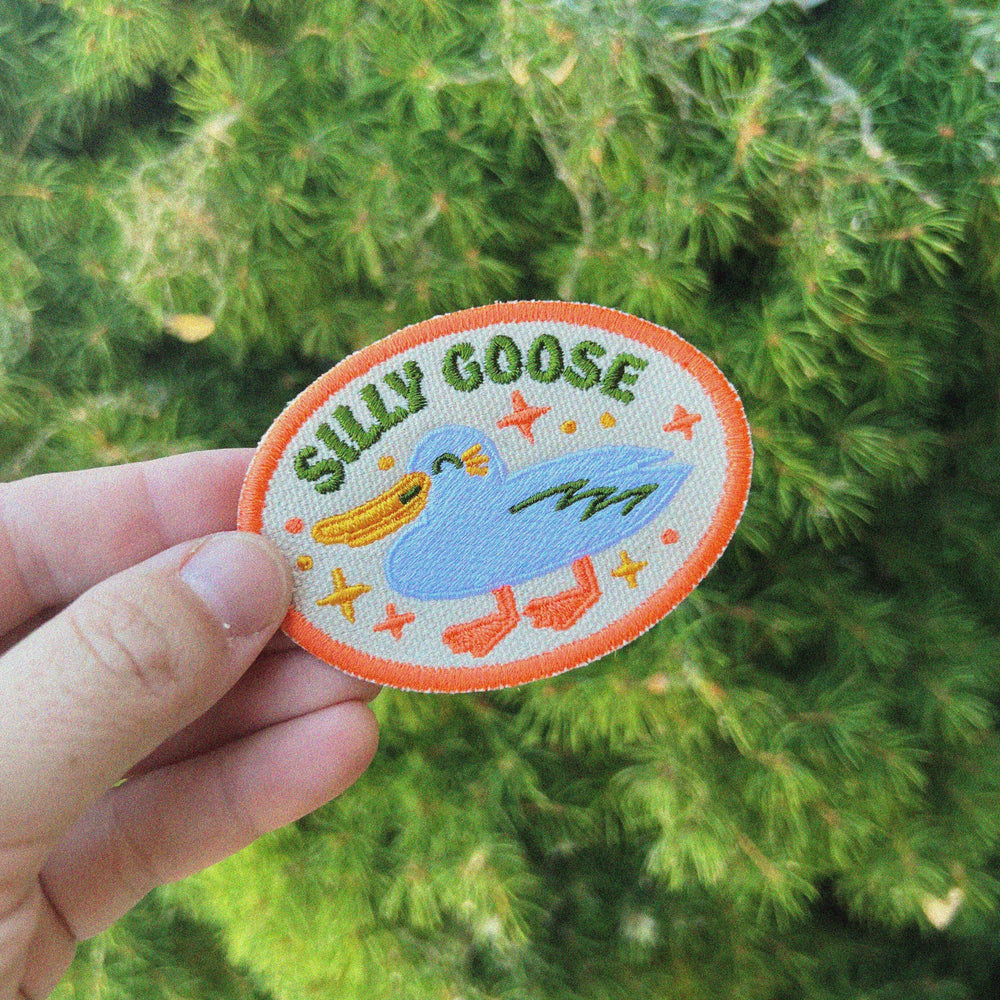 An embroidered patch being held in front of a green tree. The patch depicts a cute, white goose, winking and smiling. The style is minimalist. There are stars around it with arcing text above, reading "SILLY GOOSE." Embroidered on natural canvas.